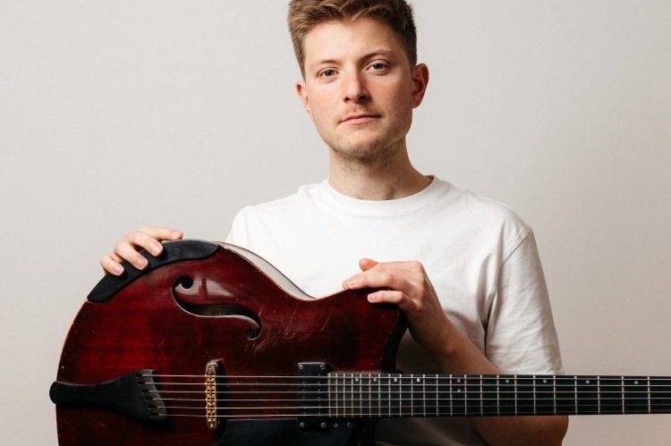 A portrait photograph of Tom Ollendorff wearing a white T-shirt and holding his guitar.