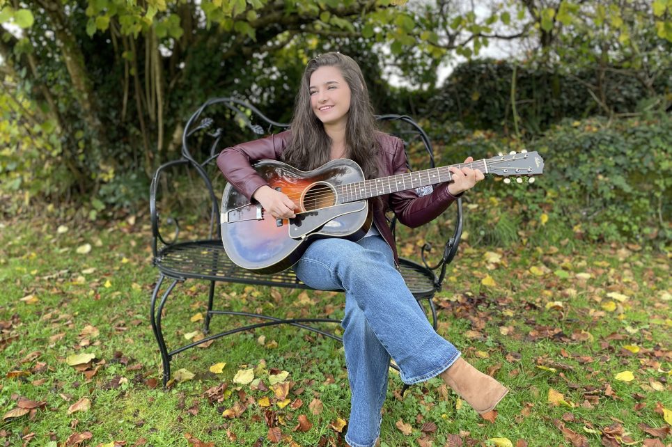 Performer Muireann Bradley sitting on a park bench strumming a guitar