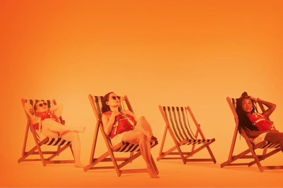 Three girls sunbathing on deck chairs on a blank orange background
