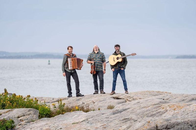 A photograph of the members of Genticorum standing on a beach with their instruments