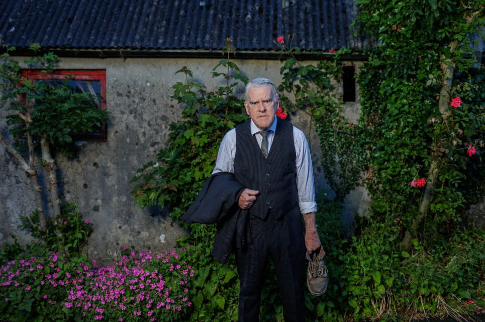 Performer Mikel Murfi wearing a suit andstanding next to a rundown cottage