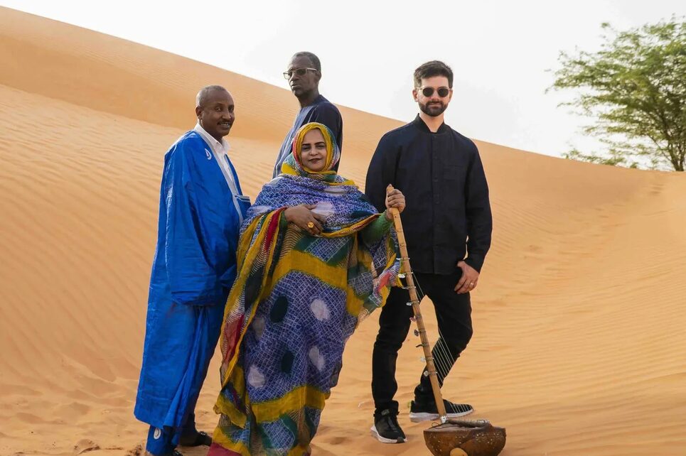 A group of four musicians in a vibrant patterned wrap, poses on a sprawling orange sand dune with a traditional stringed instrument.