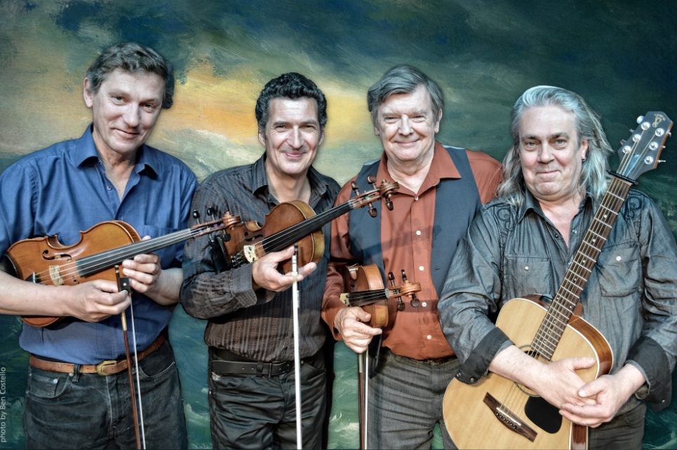 A photograph of four musicians. On the right is a player holding a guitar. The other three musicians are holding violins.