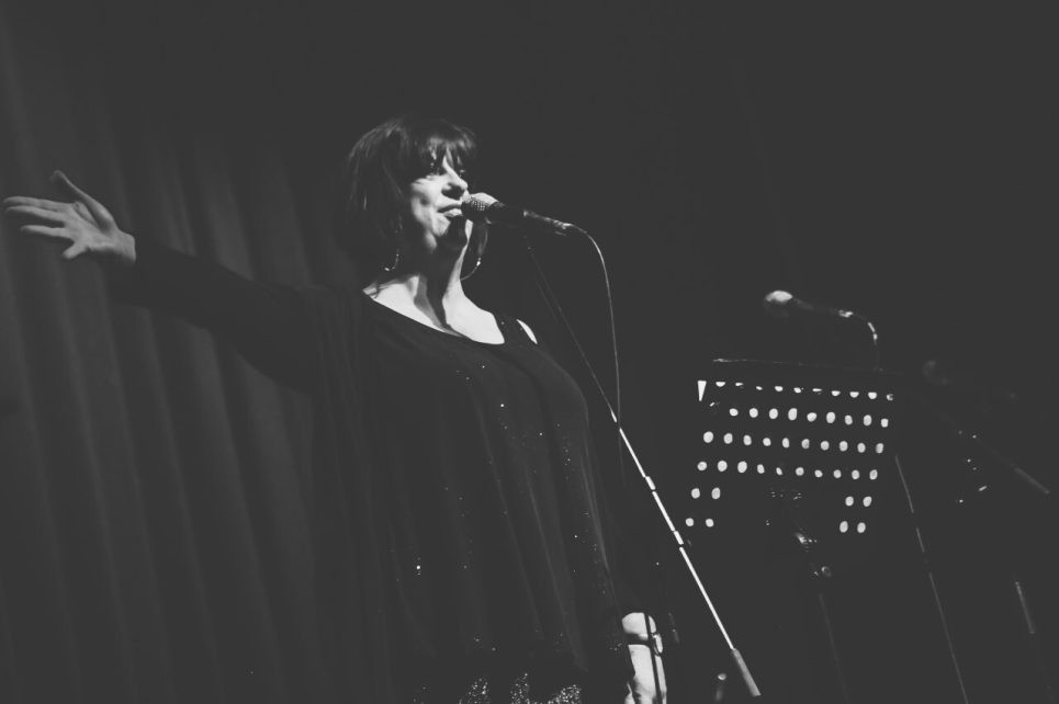 A photograph of Liane Carroll on stage, singing, her right hand outstretched.