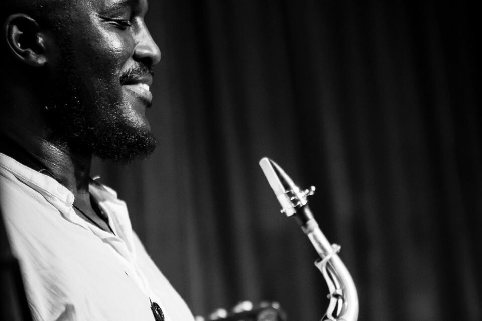 A black and white photo of Tony Kofi smiling and holding his saxophone