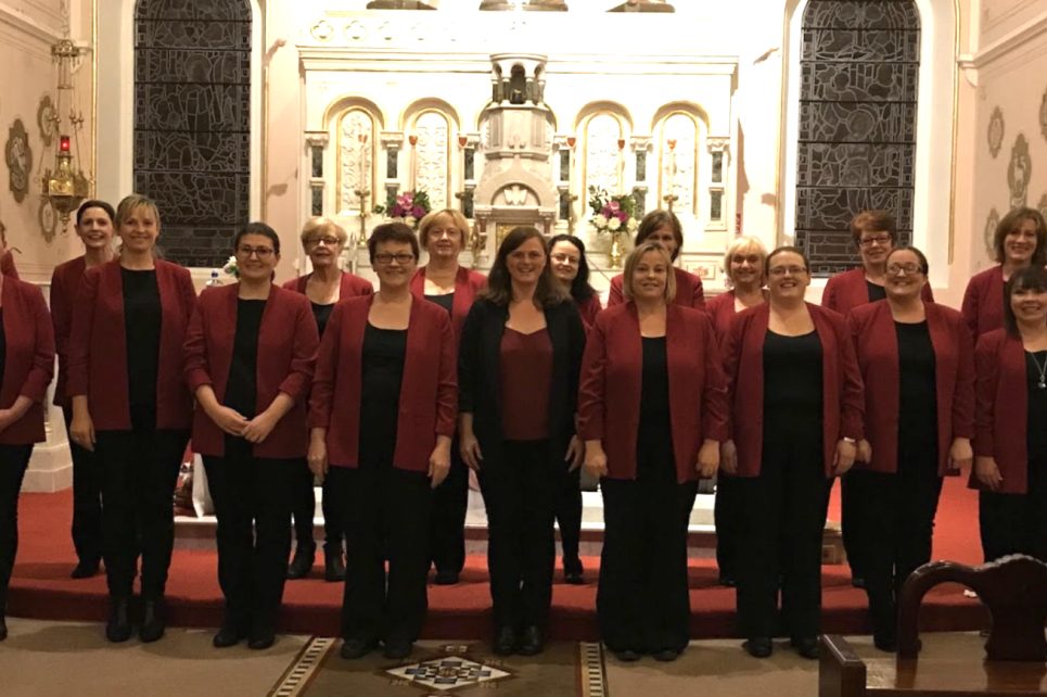 A photograph of the Coolaney Gospel Choir. They are all dressed in black pants and shirts, with dark red suit jackets over their shirts. They are standing in front of the altar in a church. A photograph of the Coolaney Gospel Choir. They are all dressed in black pants and shirts, with dark red suit jackets over their shirts. They are standing in front of the altar in a church.
