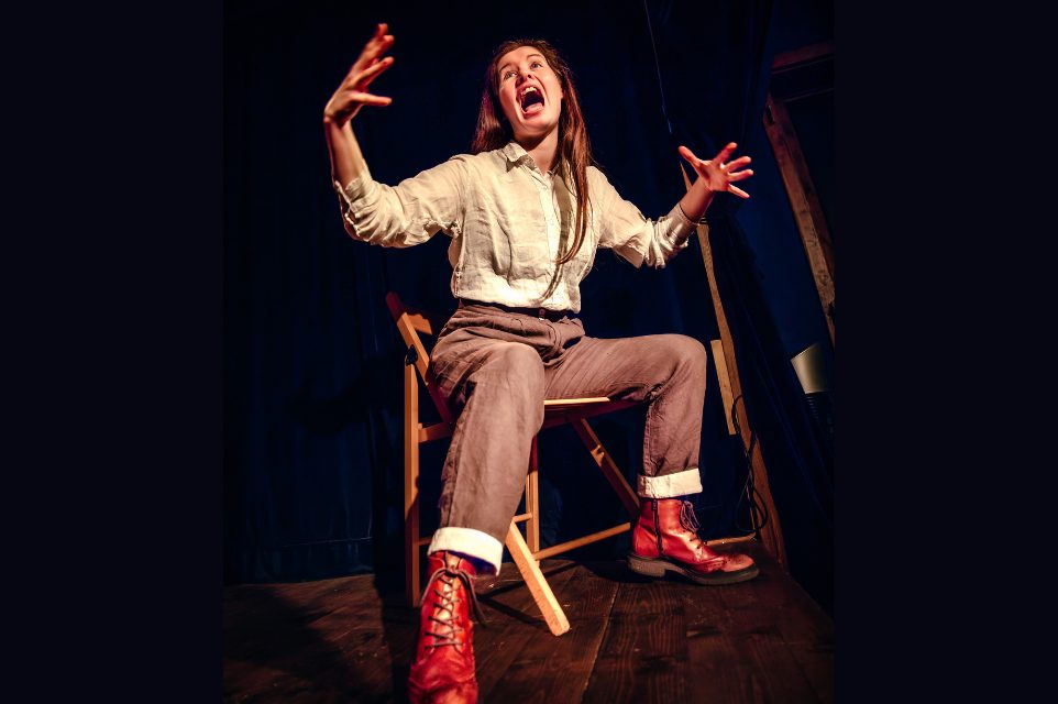 Female performer sitting on a chair yelling and with her hands raised