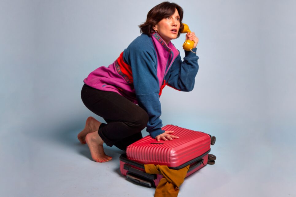 A woman in a colorful fleece jacket crouches over an overstuffed red suitcase while looking startled and holding a vintage yellow telephone receiver