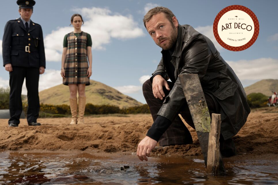 A man in a leather jacket kneels by a shoreline, reaching toward the water while a police officer and a young woman watch from behind.