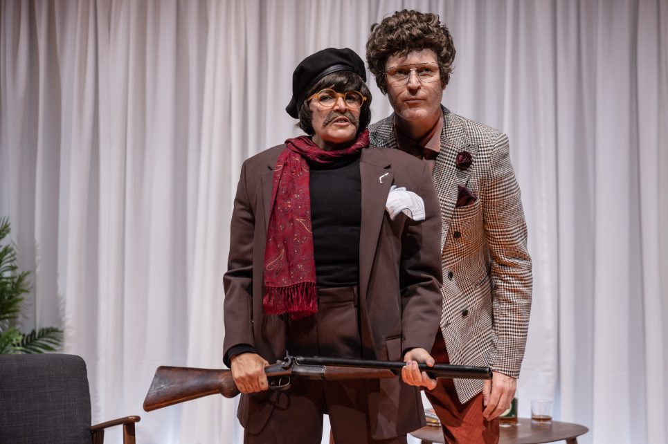 A photograph of two actors in stage. In the front is a woman wearing a wig, a fake moustache, glasses and a suit jacket. She's holding a gun. Looking over her shoulder is a man wearing a curly wig and a patterned suit jacket.