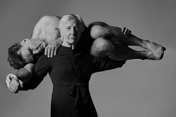 A black and white photograph depicting the dance artist Finola Cronin dressed in black and her arms spread out. Across her arms and shoulders is Mikel Murfi, who is laying on his side and curled around her. A black and white photograph depicting the dance artist Finola Cronin dressed in black and her arms spread out. Across her arms and shoulders is Mikel Murfi, who is laying on his side and curled around her.