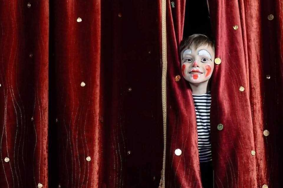 Boy in clown make-up peaking through red curtains 