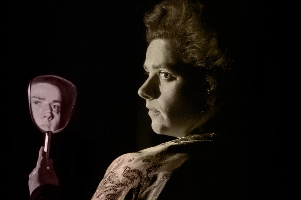 A profile photograph of Sinead Sexton in costume from her play Gazelle. She's holding up a mirror that reflects her face. A profile photograph of Sinead Sexton in costume from her play Gazelle. She's holding up a mirror that reflects her face.