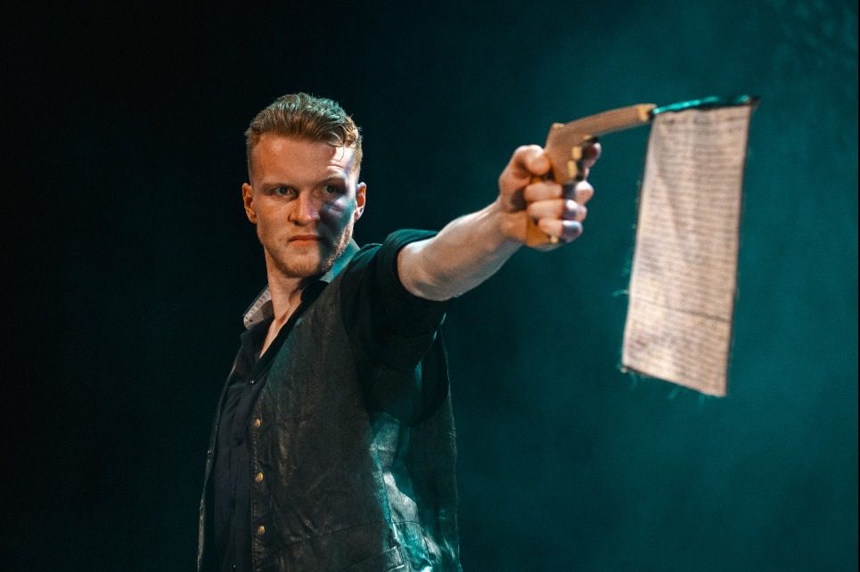 A photograph of an actor holding a gun in his outstretched left arm. A white flag is hanging out of the gun's barrel.