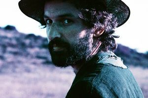 A photograph of Joachim Cooder wearing a hat and looking over his shoulder towards the camera.