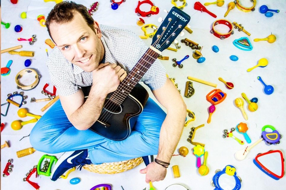A photo of the artist taking from above with him holding a guitar in his lap while sitting on the ground surrounded by a variety of colourful percussion instruments for kids A photo of the artist taking from above with him holding a guitar in his lap while sitting on the ground surrounded by a variety of colourful percussion instruments for kids