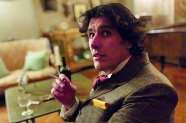 A photographs of Will Doran in character as Oscar Wilde. He's sitting in a lounge room, looking towards the camera, and holding a cigarette. A photographs of Will Doran in character as Oscar Wilde. He's sitting in a lounge room, looking towards the camera, and holding a cigarette.
