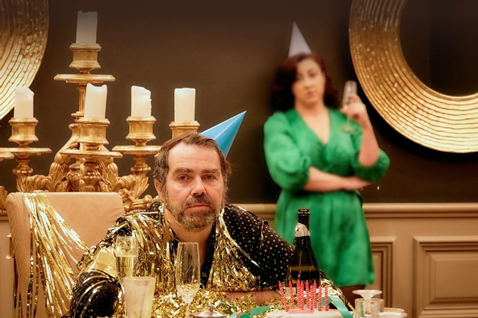A scene from Jilly Morgan's Birthday party featuring Patrick Ryan sitting at a decorated table and Georgina Miller standing in the background wearing a green dress.