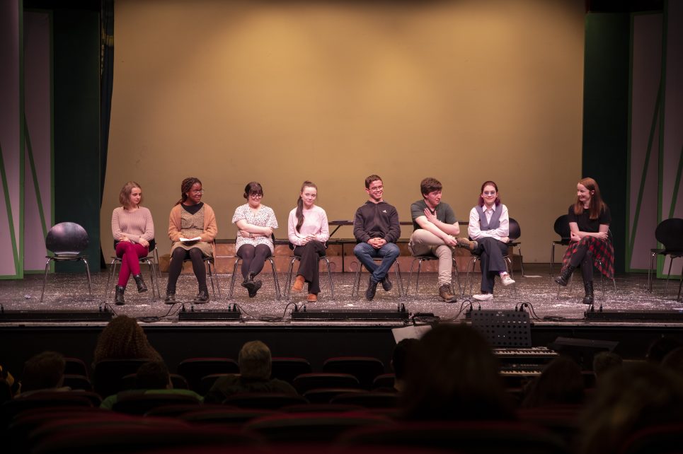 A photograph of the participants of the New Voices workshop on the Hawk's Well Theatre stage and their tutor Miriam Needham on the right hand side. A photograph of the participants of the New Voices workshop on the Hawk's Well Theatre stage and their tutor Miriam Needham on the right hand side.