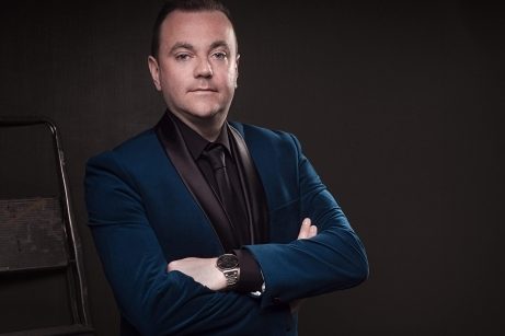 A portrait photograph of Patrick Feeney. He is wearing a black shirt and tie and a dark blue suit jacket. He is looking at the camera and his arms are folded across his chest.
