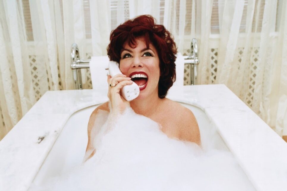 A woman with red hair laughing on a telephone while sitting in a bubble bath.