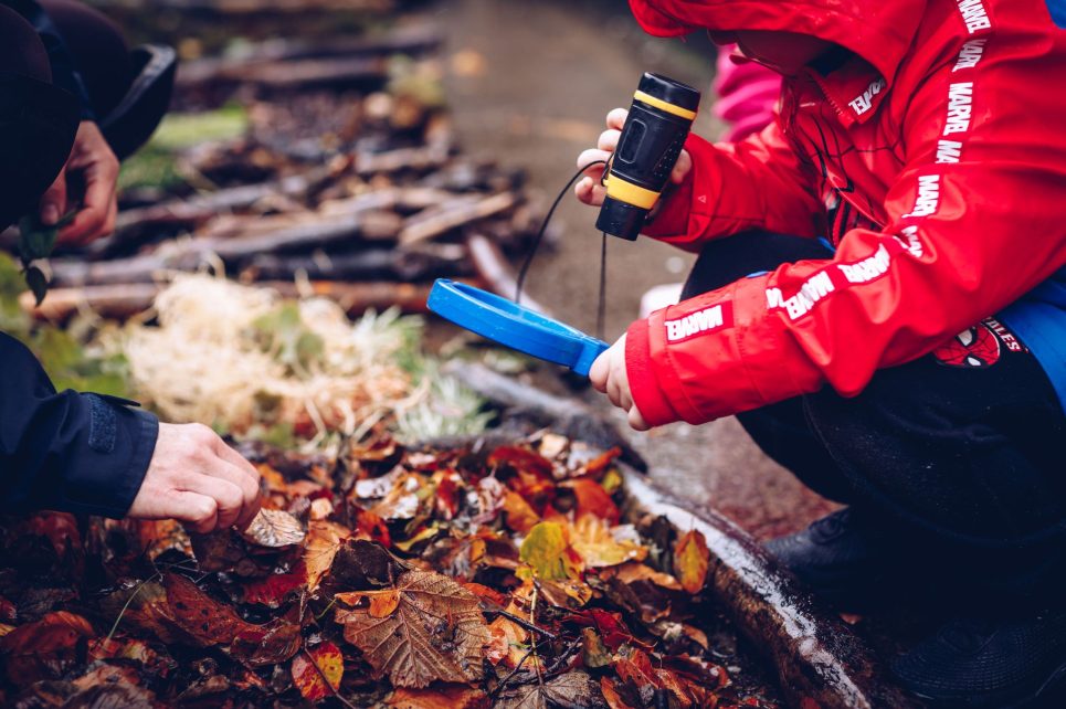 Child wearing a red raincoat holding a magnifying glass to some soil and leaves on the ground 
