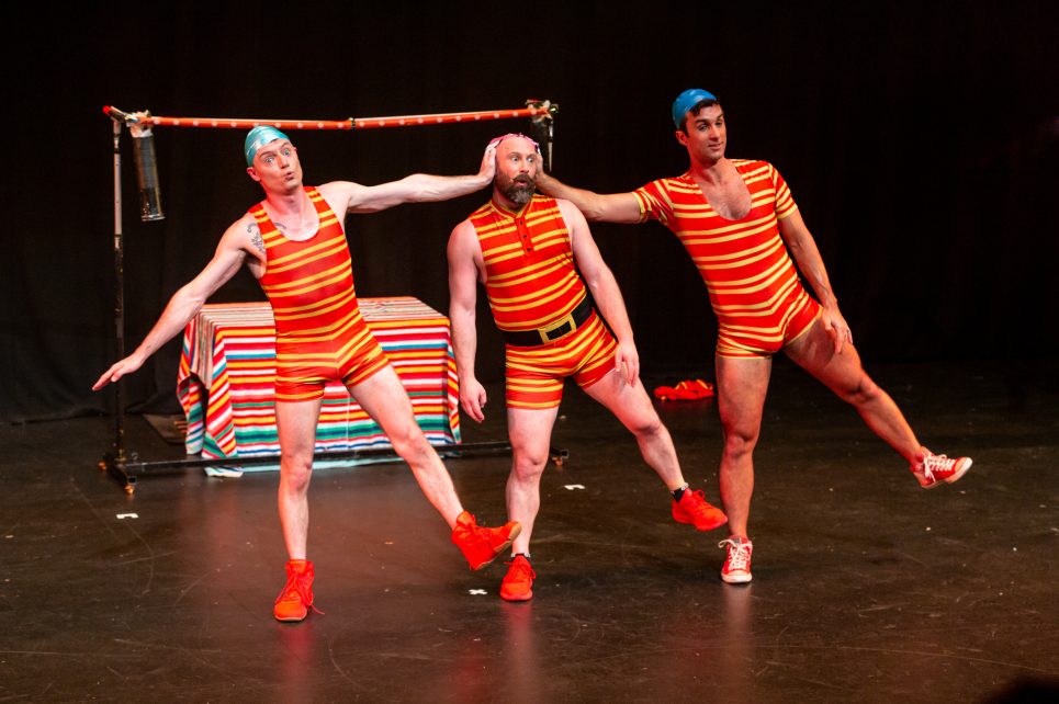A photo from the Splash Test Dummies performance, showing three actors in swimming suits and swim caps, all tilting sideways with one foot off the ground.