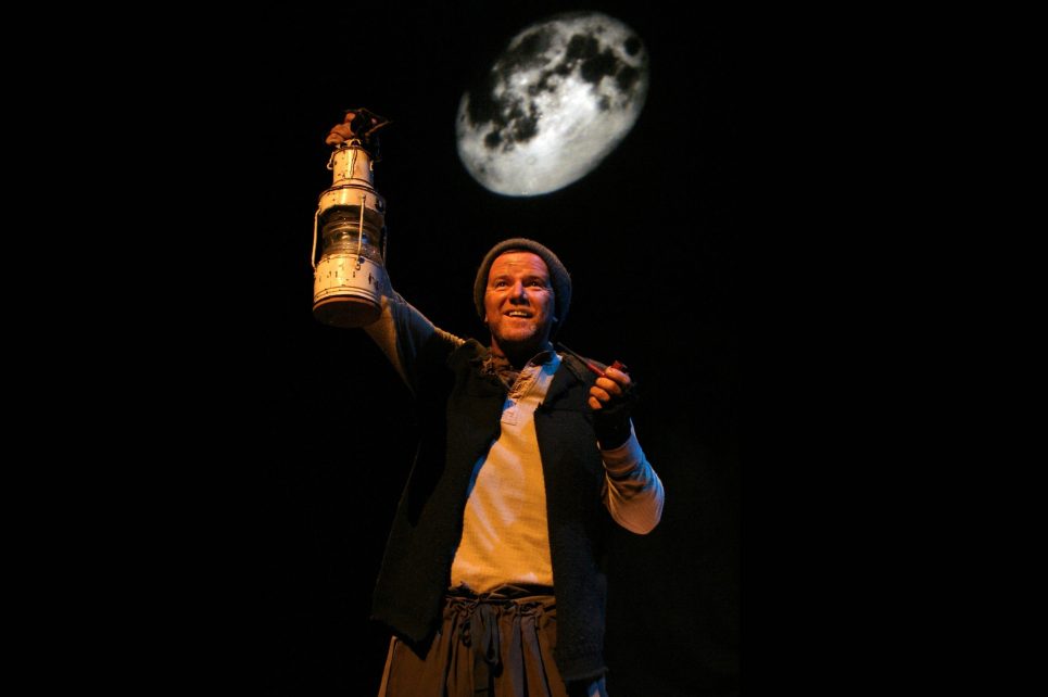 Aidan Dooley stands on stage, holding an oil lamp.