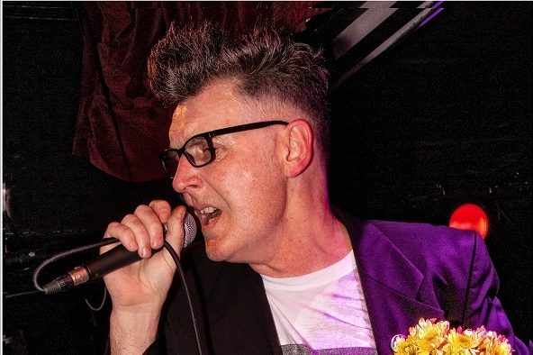 A photograph of a man in a purple jacket singing and holding flowers