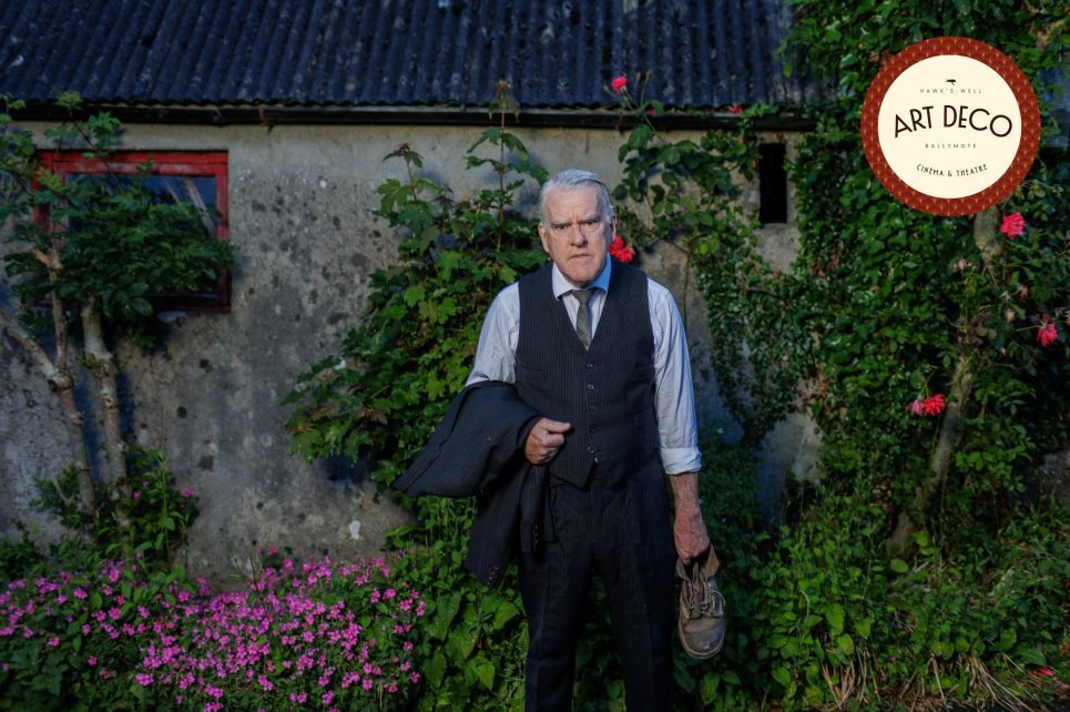 Mikel Murfi, in a vest and tie, stands outside a stone building with flowers around.
