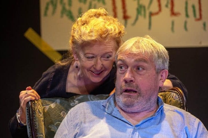 A photograph of two actors: A man sitting in a chair and looking shocked and a woman leaning over his shoulder, smiling. A photograph of two actors: A man sitting in a chair and looking shocked and a woman leaning over his shoulder, smiling.