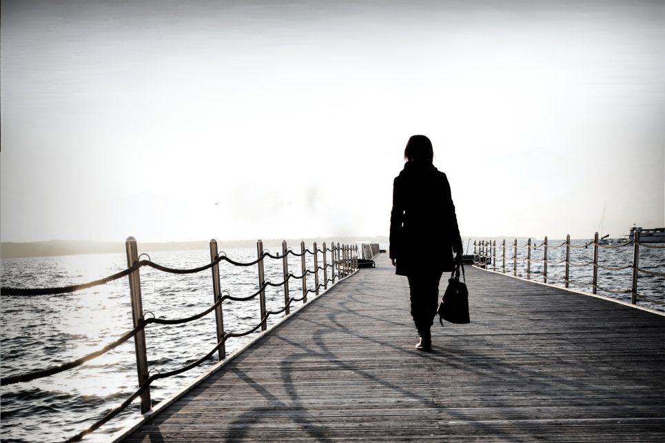 A black and white photograph of a woman walking down a pier on water