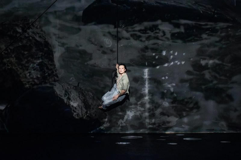 A photograph of an artist holding on to a rope and climbing up a wall