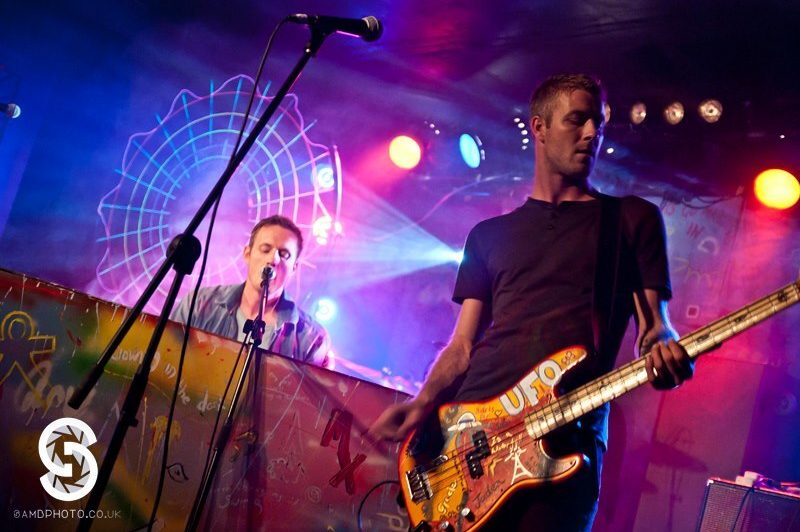 A photograph of two men performing on stage. On the left is a man with a guitar, behind him a man at a piano.