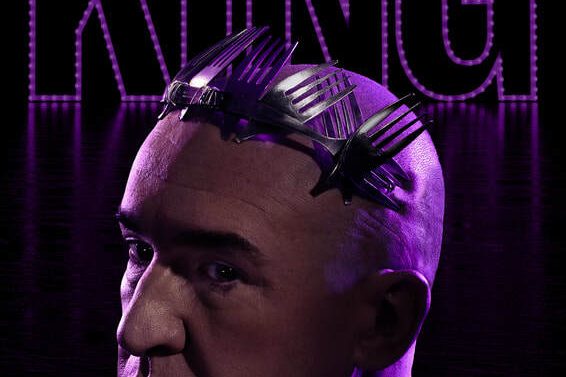 A photo of Pat Kinevane from the play King. Pat is looking to the side of the camera and is wearing a crown made out of fork tines. Above him, the word king in large letters.