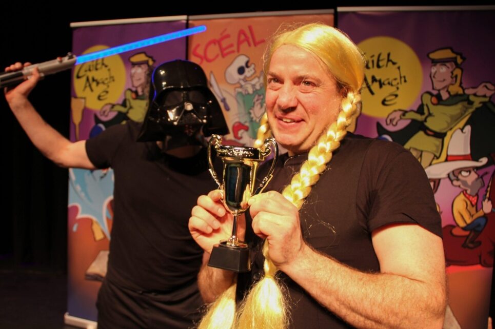In the foreground is a man wearing a blonde wig with braided pigtails. To his left is a man wearing a Darth Vader mask