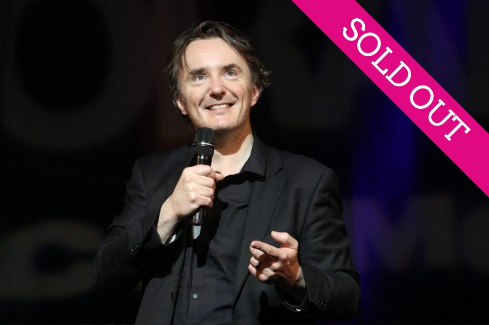 Comedian Dylan Moran standing on stage holding a microphone