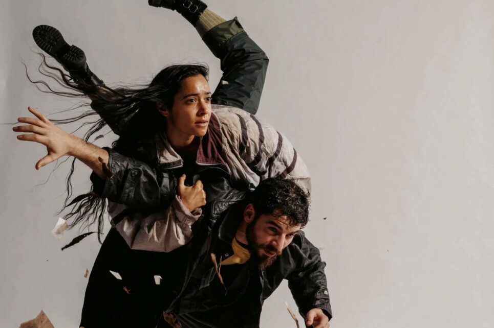 Two people in a dynamic, acrobatic pose amidst swirling dry leaves and paper.