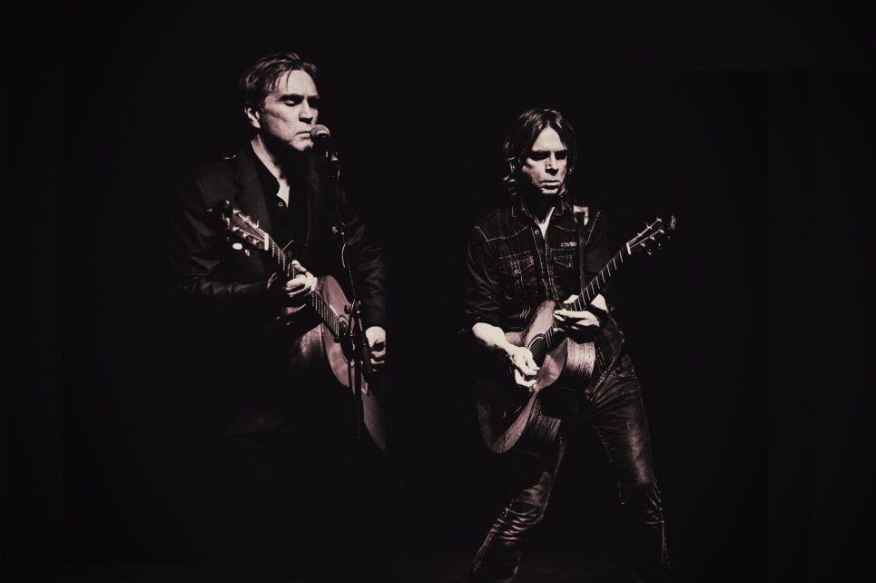 Monochrome photo of two men playing guitar posed on a stage with a microphone